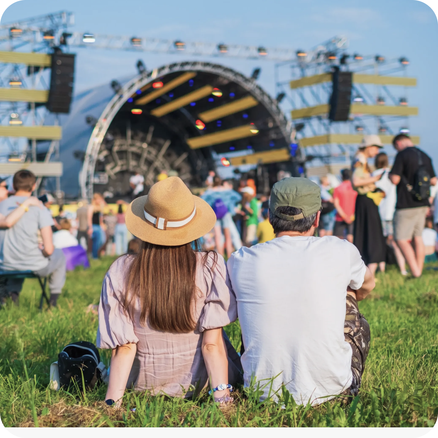 Two people sitting on grass at a music festival, seen from behind, with a large stage and crowd in the background under a blue sky.