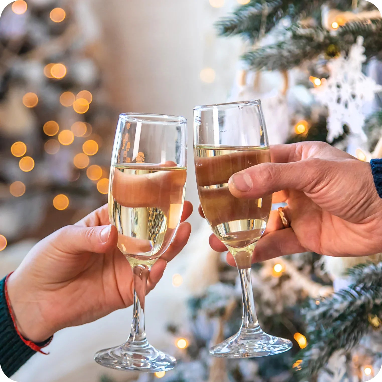 Two people clinking champagne glasses by a decorated Christmas tree with festive lights
