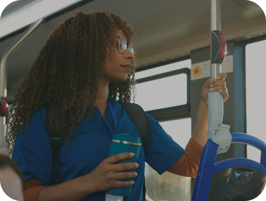 A woman with curly hair and glasses, wearing a blue scrub top, stands on a bus holding a vertical handrail and a teal travel tumbler.