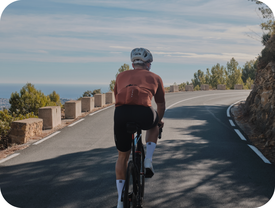 A cyclist viewed from behind riding a road bike on a winding mountain road under a blue sky, wearing a rust-orange short-sleeve cycling jersey, black shorts, white socks, and a silver helmet.
