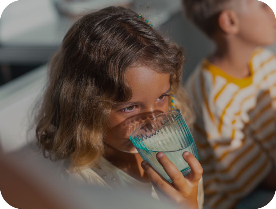 A young girl with brown hair drinks from a clear ribbed glass cup, with a blurred boy in a yellow and white striped shirt in the background.