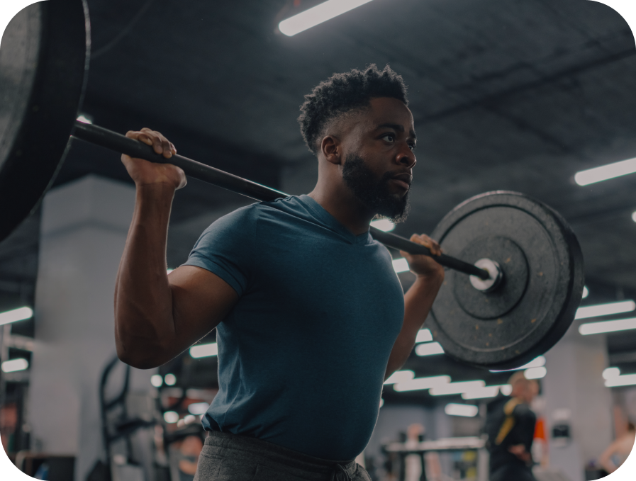 A man in a gym wearing a teal blue t-shirt and gray sweatpants, lifting a barbell with weight plates on a bench press rack.