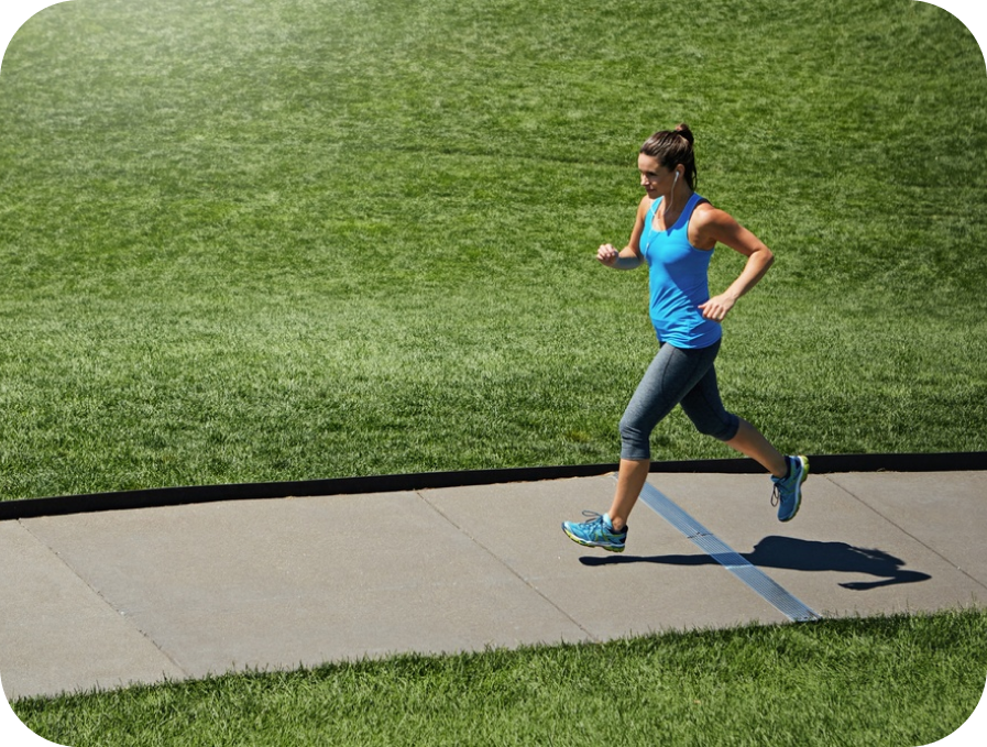 A woman jogging on a concrete path in a park, wearing a bright blue tank top, gray capri leggings, and blue running shoes, with a large green grass field behind her.