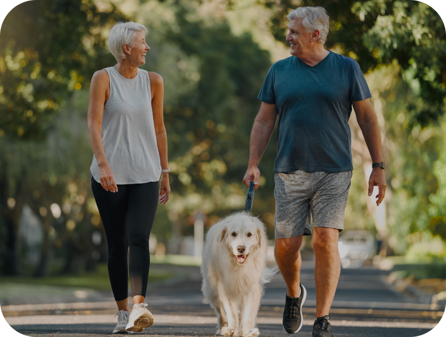 An older couple walking a fluffy white dog on a paved path outdoors, with trees and greenery blurred in the background.